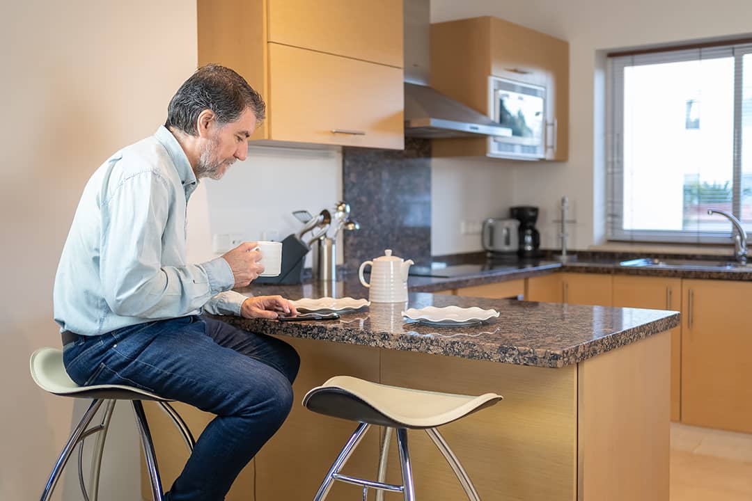 Senior man sitting on a stool in the kitchen, drinking a cup of coffee and looking at the mobile phone.